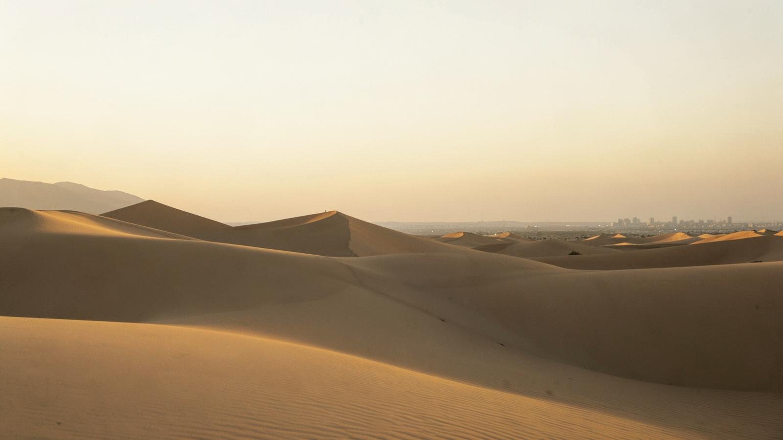 Golden desert dunes at dusk with a distant city skyline on the horizon